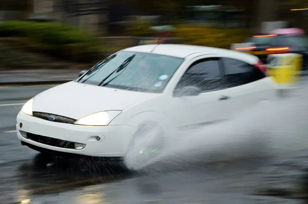 Auto bianca in movimento su strada bagnata, con un'enorme spruzzata d'acqua che indica come il fenomeno dell'aquaplaning aumenta.