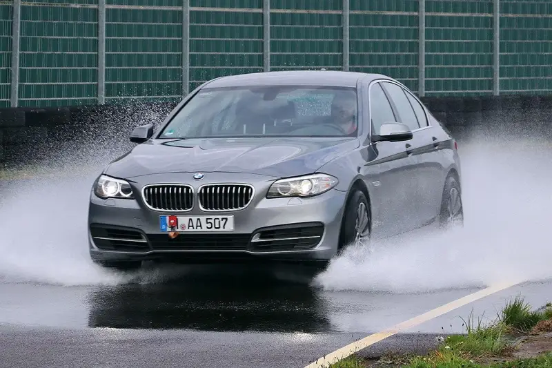 BMW grigia in test di acquaplaning, solleva un'onda d'acqua mentre sfreccia sull'asfalto bagnato.